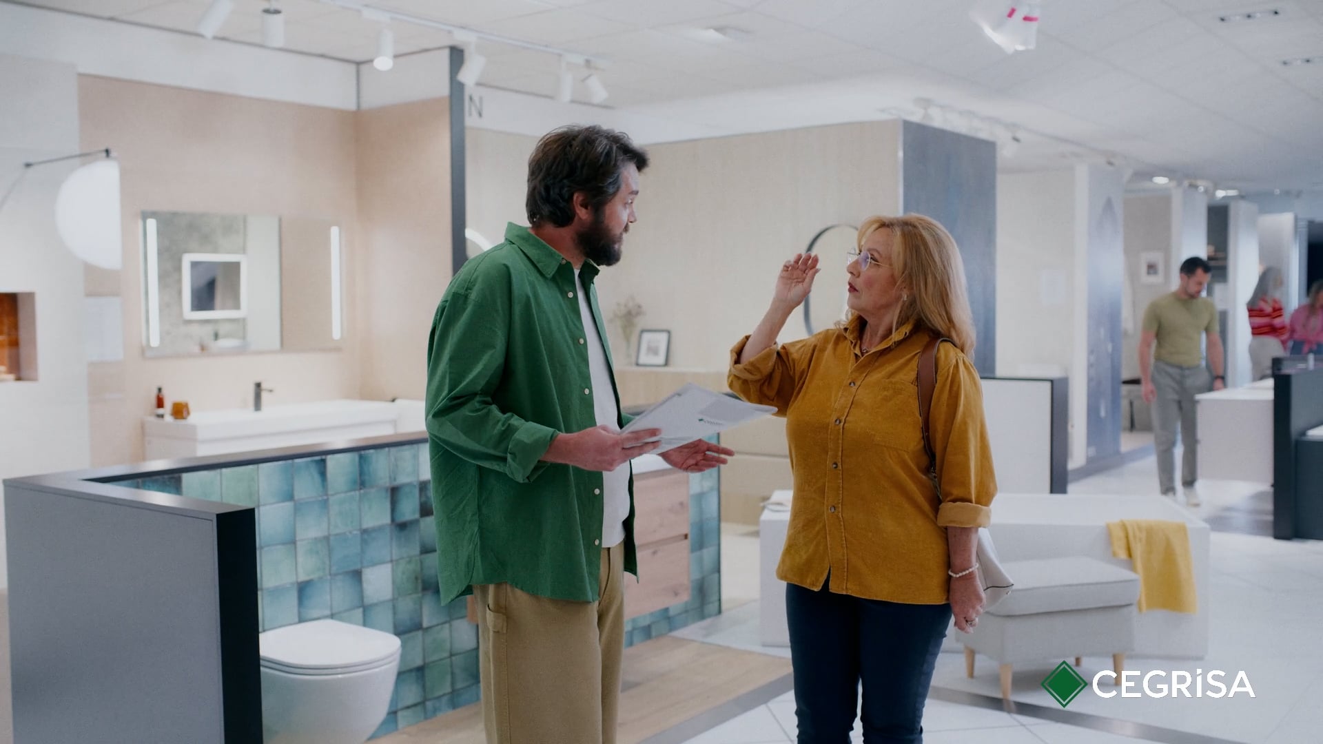 Cegrisa | Más que inspiración — Cegrisa — A male shop assistant in a green shirt talks with a middle-aged customer in a mustard-yellow shirt at a Cegrisa bathroom and kitchen showroom in the Canary Islands, with the CEGRISA logo in the corner.