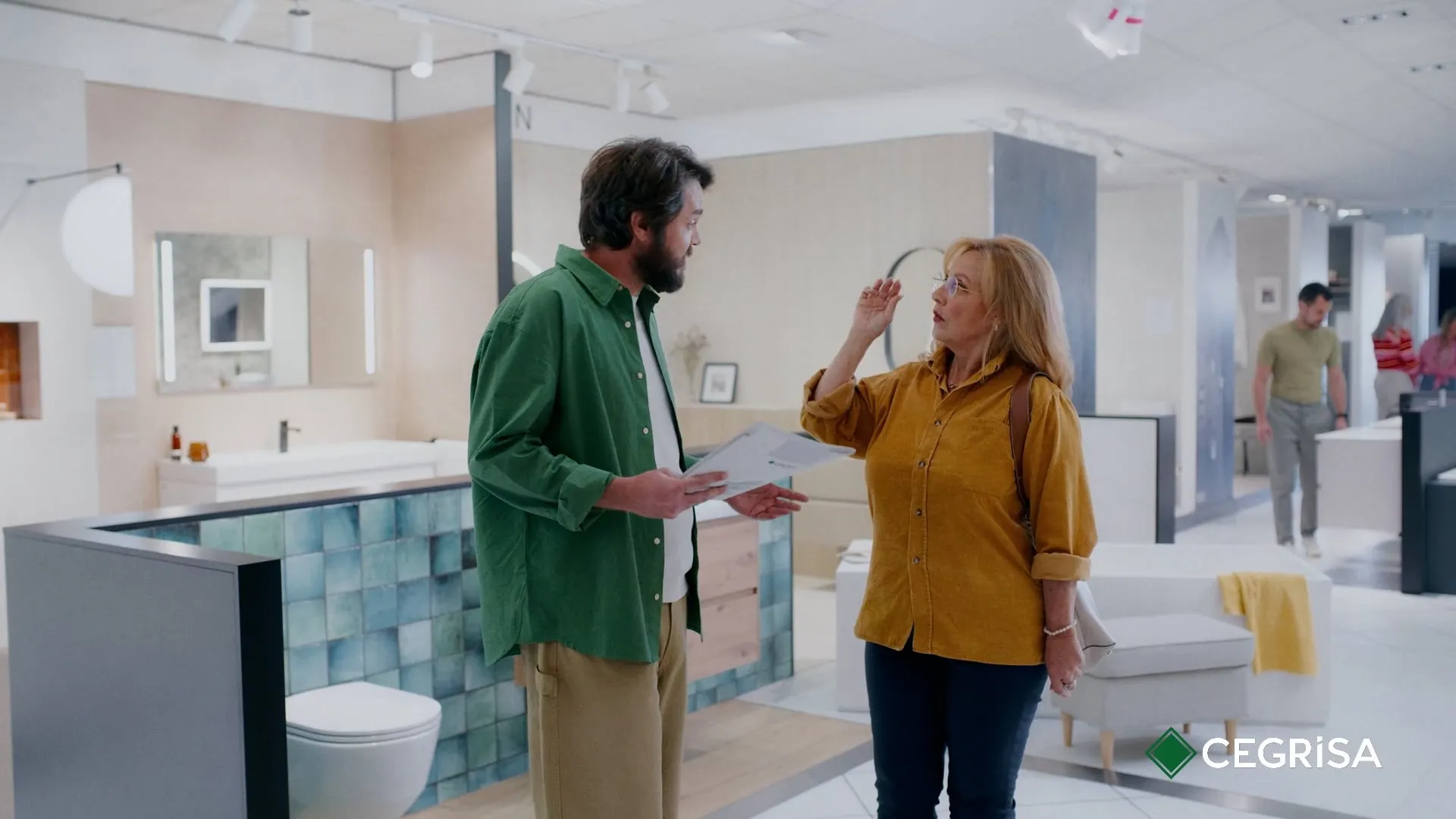 Cegrisa | Más que inspiración — Cegrisa — A male shop assistant in a green shirt talks with a middle-aged customer in a mustard-yellow shirt at a Cegrisa bathroom and kitchen showroom in the Canary Islands, with the CEGRISA logo in the corner.