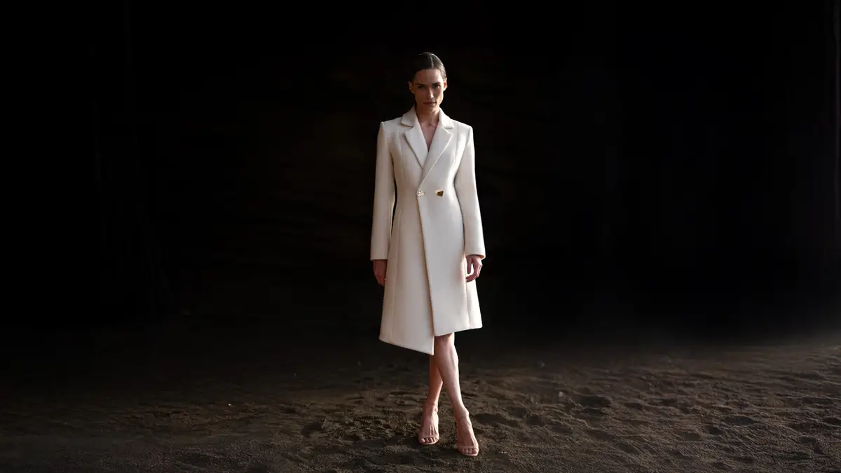 Ember by Inti — Una mujer con abrigo blanco, de pie en una playa.